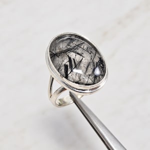 May include: A silver ring featuring a large, oval-shaped rutilated quartz gemstone. The clear stone contains distinct, needle-like black inclusions throughout its surface. The gemstone is set in a smooth, polished silver bezel with a split band design.