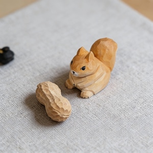 May include: A wooden squirrel ornament and a peanut. The squirrel is light brown with black eyes and a bushy tail. The peanut is also carved from wood, with detailed texture. The items are set on a neutral-coloured surface.