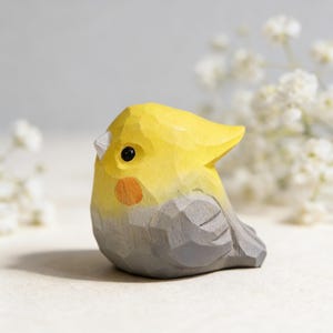 May include: A hand-carved wooden bird figurine. The bird has a yellow head and crest, a grey body, and a small orange cheek patch. The bird's eye is a small black dot. The figurine is a decorative item.
