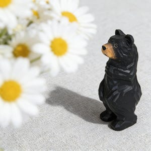 Könnte beinhalten: Eine geschnitzte Holzfigur eines Bären in schwarzer Farbe mit einem braunen Maul. Der Bär steht aufrecht vor einem neutralen Hintergrund. Weiße Gänseblümchen mit gelben Zentren sind im Hintergrund.