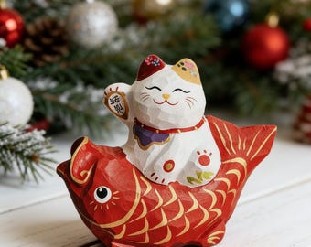 May include: A carved wooden figurine of a white cat with colorful accents riding a red fish. The cat is holding up a paw with Japanese characters. The fish has gold detailing. The background includes Christmas ornaments and greenery.