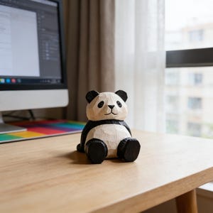 Op de afbeelding: Handgesneden houten panda beeldje met zwart-witte details. De panda zit met uitgestrekte benen. Het staat op een lichtgekleurd houten bureau. Een computermonitor en een raam zijn zichtbaar op de achtergrond.