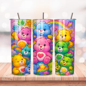 May include: Three stainless steel tumblers with colorful Care Bear designs. Each tumbler features a different arrangement of Care Bears, rainbows, and flowers. The tumblers have silver lids and straws, and are set on a wooden surface.