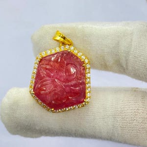 May include: A pink gemstone pendant with a floral design carved into the stone. The pendant is set in a gold frame with a halo of small, clear gemstones. The pendant is held up by a gold bail.