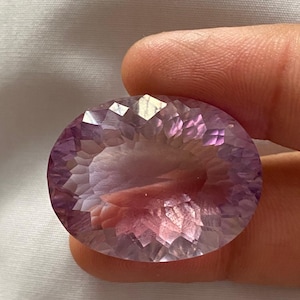 Könnte beinhalten: Ein facettierter, ovaler Amethyst-Edelstein. Der Edelstein zeigt einen Farbverlauf, der von einem tiefen Lila zu einem helleren Lavendel und Rosa übergeht. Der Schliff des Steins erzeugt mehrere reflektierende Oberflächen.