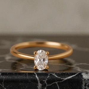 Könnte beinhalten: Ein goldener Ring mit einem klaren, oval geschliffenen Edelstein. Der Ring hat ein schlichtes, poliertes Goldband. Der Edelstein ist mit vier Krappen gefasst. Der Ring wird auf einer schwarz-grauen Marmoroberfläche präsentiert.