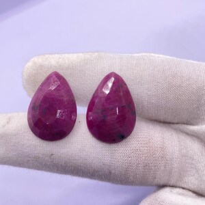 Peut inclure: Deux pierres précieuses en forme de goutte d'eau, facettées, d'un magenta profond. Les gemmes ont une surface polie avec des inclusions internes visibles. Les pierres sont présentées sur un fond de tissu blanc, mettant en valeur leur couleur et leur taille.