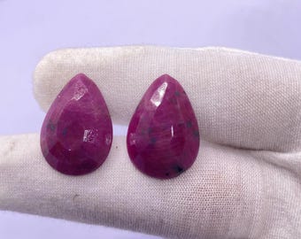 May include: Two faceted, teardrop-shaped gemstones in a deep magenta hue. The gems have a polished surface with visible internal inclusions. The stones are displayed on a white fabric background, showcasing their color and cut.
