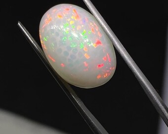 Può includere: Una gemma di opale di forma ovale, tenuta da pinzette metalliche. L'opale ha un colore di base bianco latte con lampi iridescenti di rosso, verde e giallo. La superficie della pietra presenta un sottile motivo cellulare.