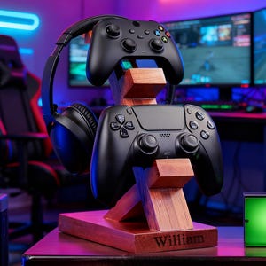 May include: A wooden gaming controller and headset stand with a personalized name. The stand holds two black controllers and a pair of black headphones. The stand is made of wood and has a natural finish. The name "William" is engraved on the base.