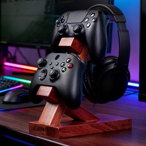 Puede incluir: Un soporte de madera para mandos de juegos y auriculares. El soporte es de madera oscura y sostiene dos mandos negros y unos auriculares negros. El soporte está sobre un escritorio de madera. La palabra "LENNY" es visible en la base.