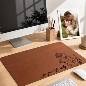 May include: A brown rectangular desk mat with a black illustration of a father and child, and the text "Love you, Daddy." A framed photo of a father and child is on the desk. A computer, plant, and desk accessories are also present.