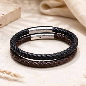 May include: Two braided leather bracelets, one black and one brown, featuring cylindrical silver-tone metal clasps. The black bracelet is engraved with the text Always & Forever xx, and the brown bracelet is engraved with I Love you and a heart symbol.