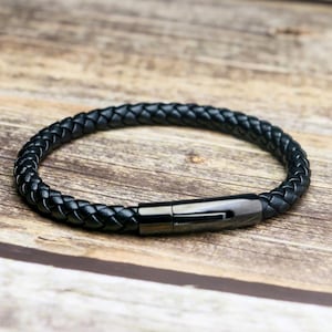 Op de afbeelding: Een zwarte gevlochten leren armband met een pistoolkleurige sluiting. De armband is cirkelvormig en ligt op een houten oppervlak. Het leer is strak geweven en de sluiting is rechthoekig.
