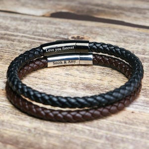 May include: Two braided leather bracelets, one black and one brown, are stacked on a wooden surface. The black bracelet has the inscription "Love you forever," and the brown bracelet says "Smith & Amy."