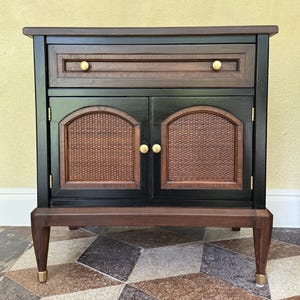 May include: A two-door cabinet with a drawer, featuring a dark wood frame and black panels. The cabinet doors have woven rattan inserts, and the drawer and doors have gold-colored knobs. The legs have gold-colored tips.