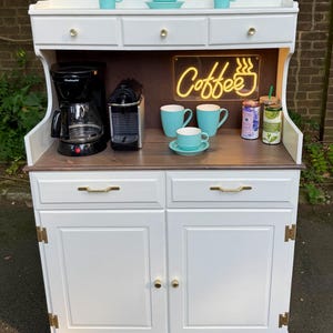 May include: White coffee station with a dark wood countertop and a white cabinet base. Features a coffee maker, espresso machine, and neon "Coffee" sign. Includes blue mugs, saucers, and decorative items. Gold hardware accents the doors and drawers.