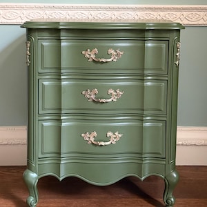 May include: A three-drawer green nightstand with ornate gold-colored handles and accents. The nightstand has a curved front and legs, and is painted in a matte finish. The nightstand is a piece of bedroom furniture.