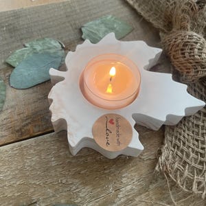 Könnte beinhalten: Ein weißer, blattförmiger Kerzenhalter mit einer brennenden Teelichtkerze. Der Kerzenhalter hat einen runden, transparenten Glaseinsatz. Ein kleiner, runder Aufkleber mit den Worten "handmade with love" befindet sich auf der Vorderseite. Der Kerzenhalter steht auf einer Holzoberfläche.