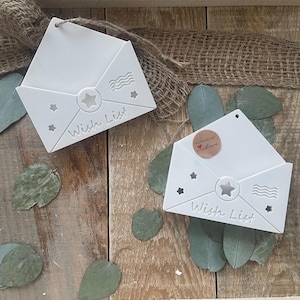 May include: Two white, envelope-shaped wall decorations with the text "Wish List" are displayed on a wooden surface. Each envelope has star and wave designs. The background includes burlap and eucalyptus leaves.