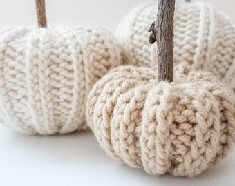 May include: Three knitted pumpkins in shades of beige and cream. The pumpkins are decorated with a twig stem.