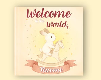 May include: A children's book cover with a soft yellow background. The cover features a white bunny riding a rocking horse. The text "Welcome to the World, Naomi" is written in burgundy and white script across the top and bottom of the cover.
