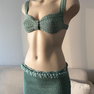 May include: A sage green two-piece crochet set consisting of a structured bralette top with wide straps and a matching midi-length skirt. The skirt features a thick, braided waistband detail, and both pieces are crafted with a ribbed knit texture.