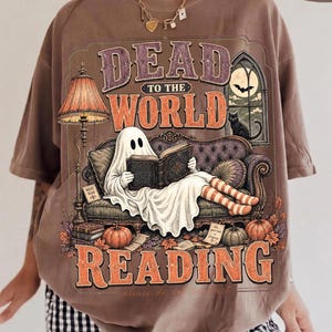 May include: An espresso-colored t-shirt featuring a graphic of a ghost sitting on a sofa reading a book. The text reads "DEAD TO THE WORLD READING". The design includes a floor lamp, a cat in a window, pumpkins, and autumn leaves in orange and brown tones.