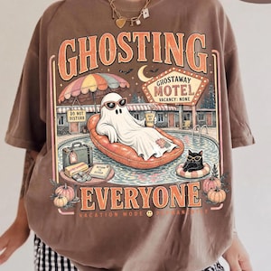 Puede incluir: Una camiseta color espresso con un gr&aacute;fico de un fantasma con gafas de sol sobre un flotador naranja. El texto dice GHOSTING EVERYONE, GHOSTAWAY MOTEL, VACANCY: NONE, DO NOT DISTURB y VACATION MODE. Tambi&eacute;n se muestran un gato negro y calabazas.