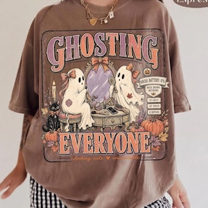 May include: An espresso-colored t-shirt featuring a graphic print with the text GHOSTING EVERYONE. The design shows two ghosts at a vanity mirror, a black cat, pumpkins, and a digital battery icon labeled SOCIAL BATTERY: 0% BUSY DOING NOTHING.