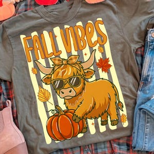 May include: A dark gray t-shirt featuring a graphic of a highland cow wearing sunglasses and a plaid bow, standing next to a pumpkin. The text "FALL VIBES" is printed in orange above the cow, set against a background of vertical cream stripes.