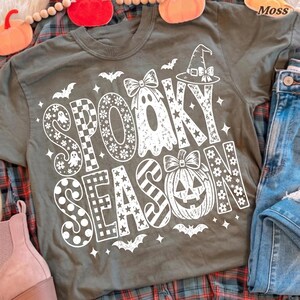 May include: An olive green t-shirt featuring white graphic text that reads SPOOKY SEASON. The design includes a ghost with a bow, a jack-o'-lantern with a bow, a witch hat, bats, and stars, with letters filled with floral, checkered, and striped patterns.