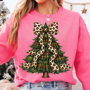 May include: A bright pink sweatshirt featuring a graphic of a green Christmas tree with a plaid pattern. The tree is adorned with a large, flowing bow that has a tan and black leopard print design. The sweatshirt has a crew neckline.