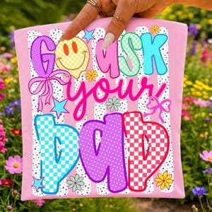 Op de afbeelding: Een lichtroze stoffen item met een grafische print met de tekst GO ASK YOUR DAD in kleurrijke, gedessineerde letters. Het ontwerp bevat een geel smileygezicht, roze strikken, groenblauwe sterren en kleine bloemen op een witte achtergrond met zwarte stippen.