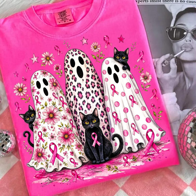 May include: A bright pink t-shirt featuring a graphic of three ghosts draped in white sheets with pink floral, leopard print, and polka dot patterns. Two black cats are integrated into the design, accented by pink awareness ribbons.