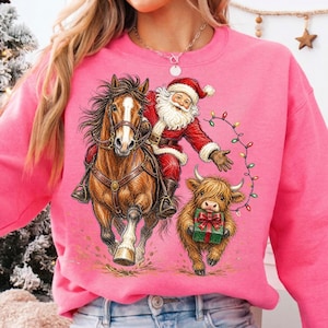 Puede incluir: Una sudadera rosa brillante con una ilustraci&oacute;n gr&aacute;fica de Santa Claus montando un caballo marr&oacute;n, acompa&ntilde;ado por una peque&ntilde;a vaca de las Tierras Altas que sostiene un regalo verde. Luces navide&ntilde;as de colores rodean las figuras.