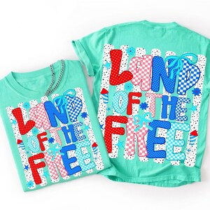 May include: Two mint green t-shirts featuring a graphic print with the text LAND OF THE FREE. The design includes red, white, and blue letters with polka dot and checkered patterns, accented by small blue flowers, stars, and firework illustrations.