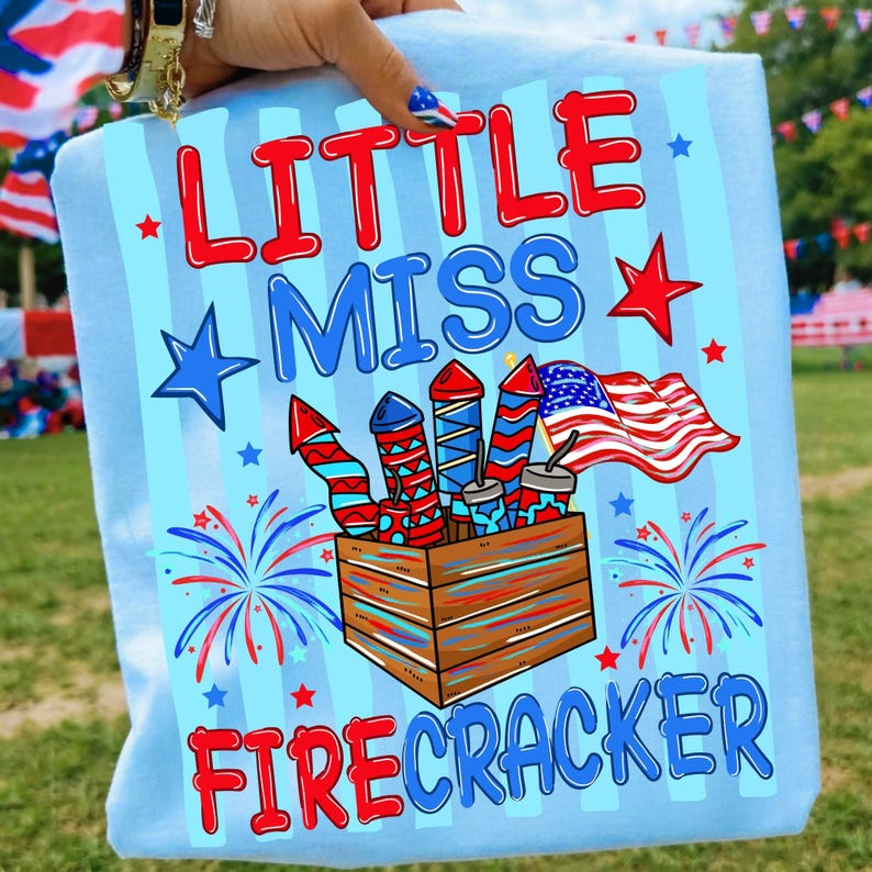 Może przedstawiać: Jasnoniebieski t-shirt z nadrukiem graficznym z napisem LITTLE MISS FIRECRACKER w kolorze czerwonym i niebieskim. Wz&oacute;r przedstawia drewnianą skrzynkę wypełnioną czerwono-biało-niebieskimi fajerwerkami, amerykańską flagę oraz kolorowe gwiazdki.