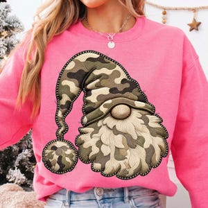 K&ouml;nnte beinhalten: Ein leuchtend pinkfarbenes Langarm-Sweatshirt mit einer gro&szlig;en, mittig aufgestickten Applikation eines Gnoms. Der Gnom tr&auml;gt eine M&uuml;tze mit Camouflage-Muster und passendem Bommel sowie einen strukturierten, cremefarbenen Bart und eine runde Nase.