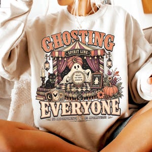 Pode incluir: Uma camisola de gola redonda cor de creme com uma impress&atilde;o gr&aacute;fica de um fantasma com turbante atr&aacute;s de uma bola de cristal. O texto diz GHOSTING EVERYONE, SPIRIT LINE, HELLO? YOU THERE? ANSWER ME, AVAILABLE GHOSTING e NO APPOINTMENTS NO APPARITIONS.