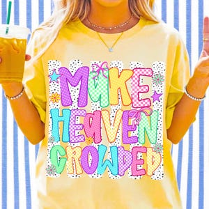 May include: A pale yellow t-shirt featuring the text "MAKE HEAVEN CROWDED" in colorful, patterned block letters. The design includes pink, blue, yellow, and green checkerboard and polka dot patterns, accented with small flowers, stars, and bows.
