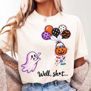 May include: A cream-colored t-shirt featuring a graphic of a white ghost reaching toward a pumpkin bucket filled with candy, lifted by five colorful balloons. The text "Well, sh*t..." is printed in black below the ghost and the pumpkin bucket.