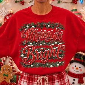 Może przedstawiać: Czerwona bluza z okrągłym dekoltem z napisem Merry & Bright stylizowaną czerwoną czcionką. Tekst otoczony jest zielonymi gałązkami sosny, białymi akcentami śniegu oraz małymi czerwonymi i białymi bombkami ułożonymi w prostokątną ramkę.