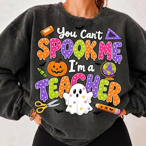 May include: A charcoal gray crewneck sweatshirt featuring a colorful graphic with the text "You Can't SPOOK ME I'm a TEACHER". The design includes a jack-o'-lantern, a ghost, a globe, scissors, a pencil, a ruler, an eraser, and small bats.
