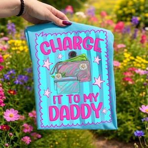 May include: A light blue t-shirt featuring a vertical striped background and a pink scalloped border. The graphic displays a wallet with green paper money, two credit cards, and white daisy flowers, with the text "CHARGE IT TO MY DADDY" in pink.
