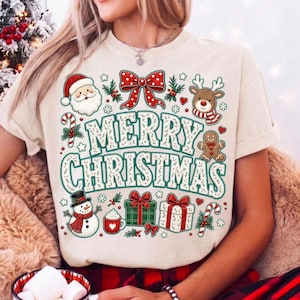 May include: A cream-colored t-shirt featuring a large graphic with the text MERRY CHRISTMAS in green and white. The design includes a Santa Claus, a reindeer, a gingerbread man, a snowman, a red bow, gift boxes, and candy canes.