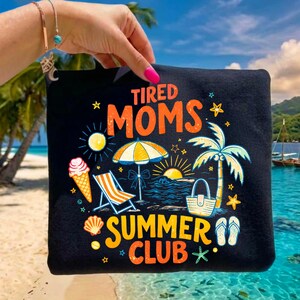 May include: Black tote bag with a colorful summer-themed graphic. The design includes the words "Tired Moms Summer Club" with illustrations of an ice cream cone, beach chair, umbrella, palm tree, and sunset.