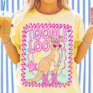 Op de afbeelding: Een lichtgeel t-shirt met een afbeelding van een kangoeroe die een roze hartvormige zonnebril en roze cowboylaarzen draagt. De kangoeroe heeft een roze strik op het hoofd. De tekst TOODLE LOO staat in grote roze letters boven de kangoeroe, omringd door een roze kronkelige rand en gele sterren.
