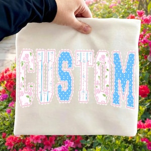 May include: Cream-colored fabric with the word "CUSTOM" in large, decorative letters. The letters have a blue and white floral pattern with pink scalloped edges. The fabric is held by a person.