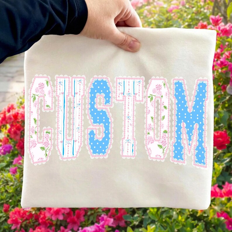 May include: Cream-colored fabric with the word "CUSTOM" in large, decorative letters. The letters have a blue and white floral pattern with pink scalloped edges. The fabric is held by a person.